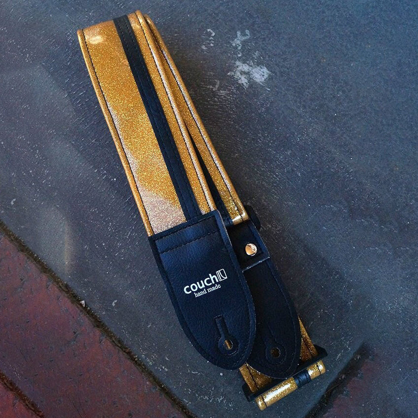 A gold sparkle guitar strap with black racing stripe, laid out on a surface.