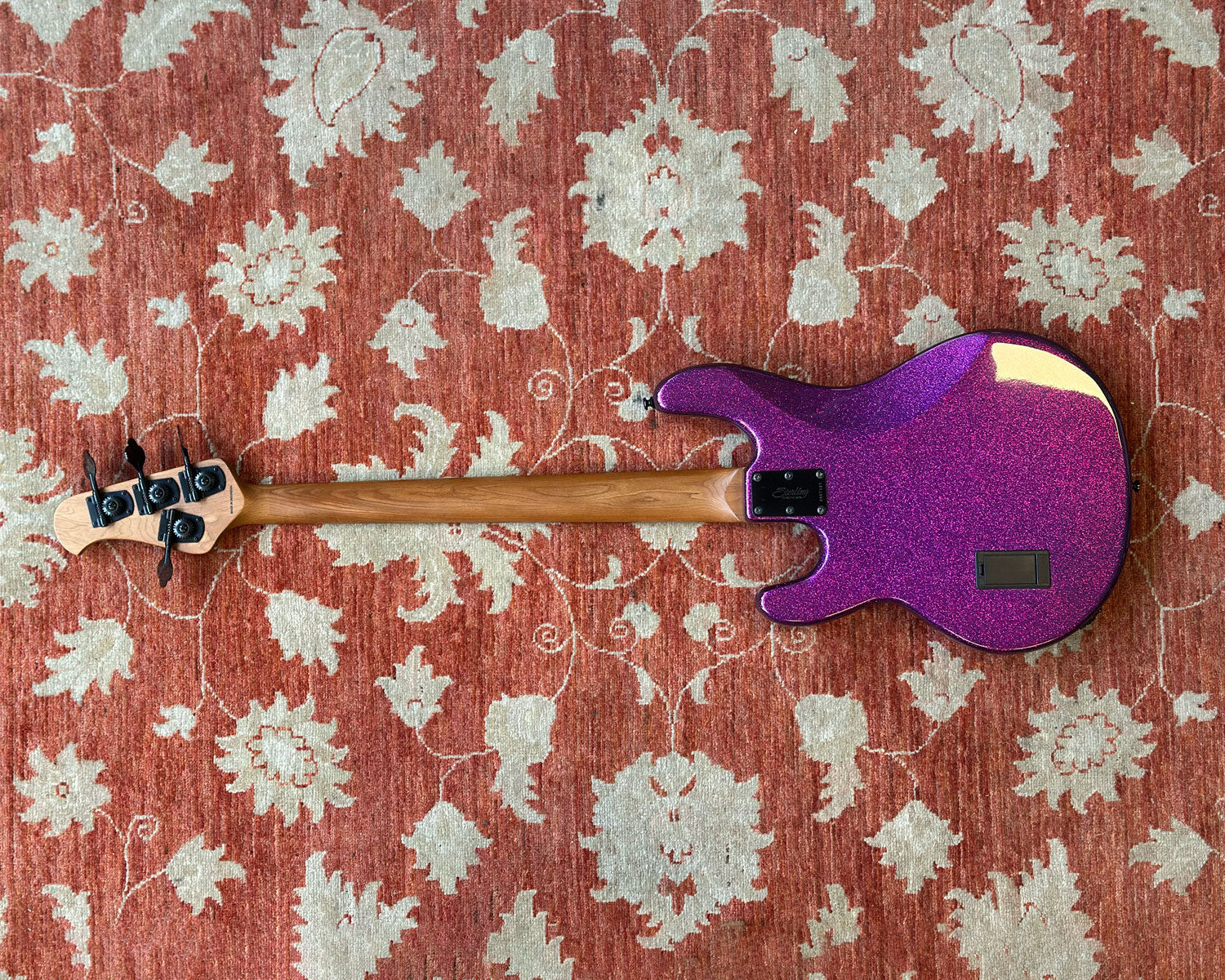 A purple glitter electric bass guitar with a natural wood neck, laid flat on a red and beige patterned rug.