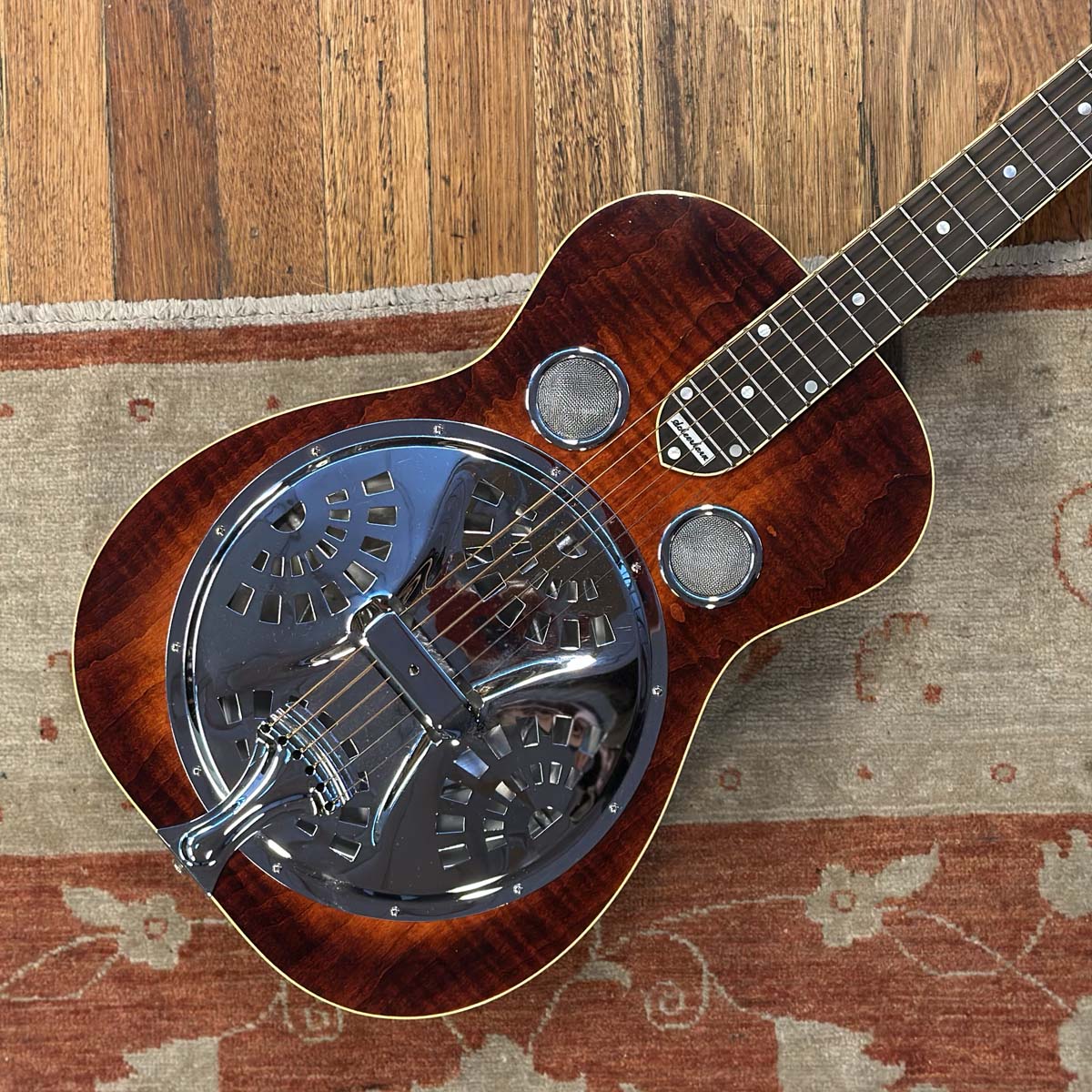 A resonator guitar with a dark reddish-brown wood body and a chrome metal resonator cavity, shown resting on a patterned rug over a wooden floor.